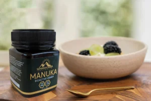 A jar of Sunhighlands Australia Manuka Bioactive Honey (MGO 550+, KPA 15.6) sits on a wooden cutting board next to a bowl of fresh fruit (blackberries and green grapes) and a golden spoon, ready for a healthy meal or snack.