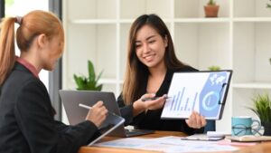 Two businesswomen discussing data while reviewing charts on a digital tablet in a modern office.